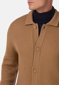 Bruine geribbelde gebreide cardigan met een kraagontwerp, voorzien van twee knopen en een gestructureerd oppervlak. Draagt over een donkere coltrui.