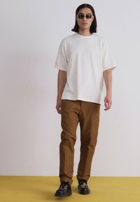 Witte oversized t-shirt, bruine tapered broek en zwarte patroon schoenen. Accessoires omvatten zwarte zonnebril. Eenvoudige achtergrond en gele vloer.