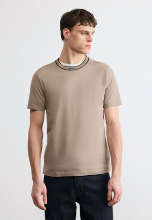 Jeune homme portant un t-shirt beige à manches courtes avec "BOSS" sur le col, jean foncé, regardant sur le côté, sur un fond uni clair.