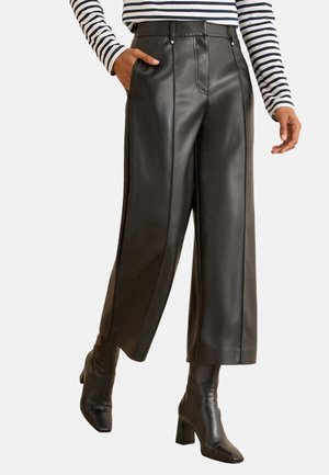 Personne portant un pantalon en cuir noir à jambes larges, une chemise à manches longues rayée et des bottines noires à talons, une main dans la poche.