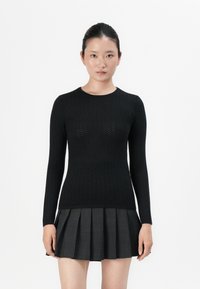 Ženska s temnimi lasmi v eleganten šopku, oblečena v prilegajočo črno teksturirano pulover in temno nabrano krilo, stoji ob belem ozadju.