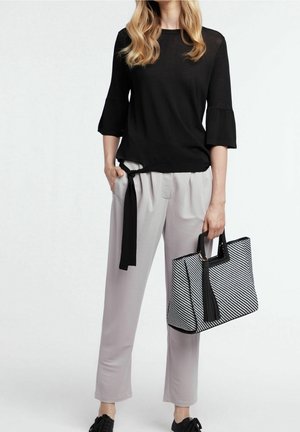 Femme portant un haut noir, un pantalon gris clair avec un détail de ceinture noire, des chaussures noires, et tenant un sac à main rayé noir et blanc.