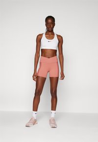 Fehér sportmellény fekete Nike logóval, rózsaszín magas derekú rövidnadrággal párosítva. A modellen világos rózsaszín sportcipők és fehér zoknik vannak.