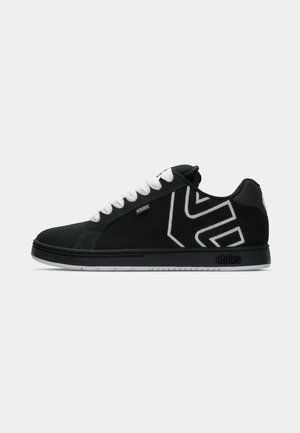 Zapatilla negra de corte bajo con cordones blancos, logo geométrico blanco en el lateral, etiqueta "etnies" y suela negra con la marca "etnies".