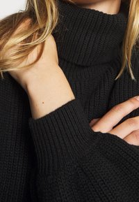 Gros plan d'une personne à la peau claire et aux cheveux blonds portant un pull à col roulé en tricot noir, la main reposant près du cou.