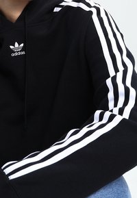 Black hoodie featuring a front pocket and three white stripes along the sleeves. It has a small white Adidas logo on the chest.