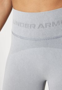 Svetlo modre športne legice z teksturiranim pasom, na katerem je v kontrastni barvi napis "UNDER ARMOUR". Gladka površina tkanine.