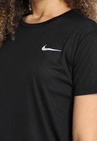 Černé sportovní tričko s krátkým rukávem, kulatým výstřihem a jemnou texturou. Na levé straně hrudi se nachází malé bílé logo Nike.