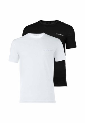 Giovane uomo con capelli biondi che indossa una maglietta nera Emporio Armani e pantaloni neri, in piedi e rivolto in avanti su sfondo bianco.