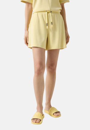 Osoba ubrana w bladoróżowe szorty ze ściągaczem i dopasowane klapki, stojąca na tle jednolitego, jasnego tła.