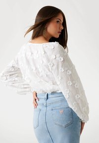 Blusa bianca con motivi floreali ricamati, maniche ampie, abbinata a jeans di denim azzurro chiaro con un logo sulla tasca posteriore.