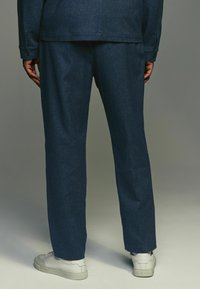 Pantaloni in denim blu scuro con taglio dritto, caratterizzati da un tessuto testurizzato. Indossati con sneaker bianche, che evidenziano linee pulite e senza branding visibile.