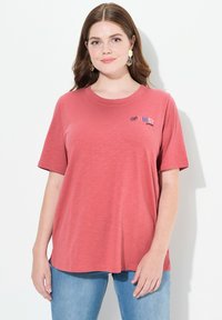 T-shirt de manga corta color coral con cuello redondo y corte relajado. Presenta el texto "OPTIMIST by PURE" en colores contrastantes en la parte delantera.