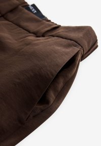 Pantaloni di cotone marrone con una texture liscia, caratterizzati da una cintura in vita e una tasca laterale, con orlo piegato per un aspetto casual.