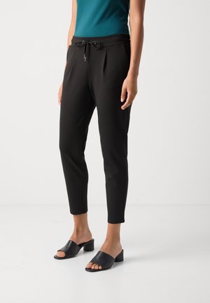 Žena stojící na bílé podlaze, oblečená v černých zúžených kalhotách se stahovací šňůrkou, tyrkysovém topu a černých mulech s otevřenou špičkou a blokovým podpatkem.