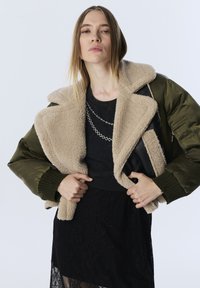 Eine Frau trägt eine grüne Bomberjacke mit beigem Sherpa-Futter über einem schwarzen Spitzenkleid und geschichteten silbernen Halsketten vor einem schlichten Hintergrund.