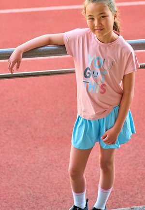 Ragazza giovane con abbigliamento sportivo appoggiata a una ringhiera di metallo sulla pista, indossa una maglietta rosa con la scritta "You Got This" e pantaloncini blu.