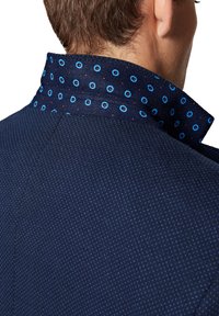 Granatowa kurtka z teksturowanej tkaniny z kołnierzykiem w grochy w odcieniach niebieskiego i czerwonego, ukazująca wyraźne wzory i kontrasty.