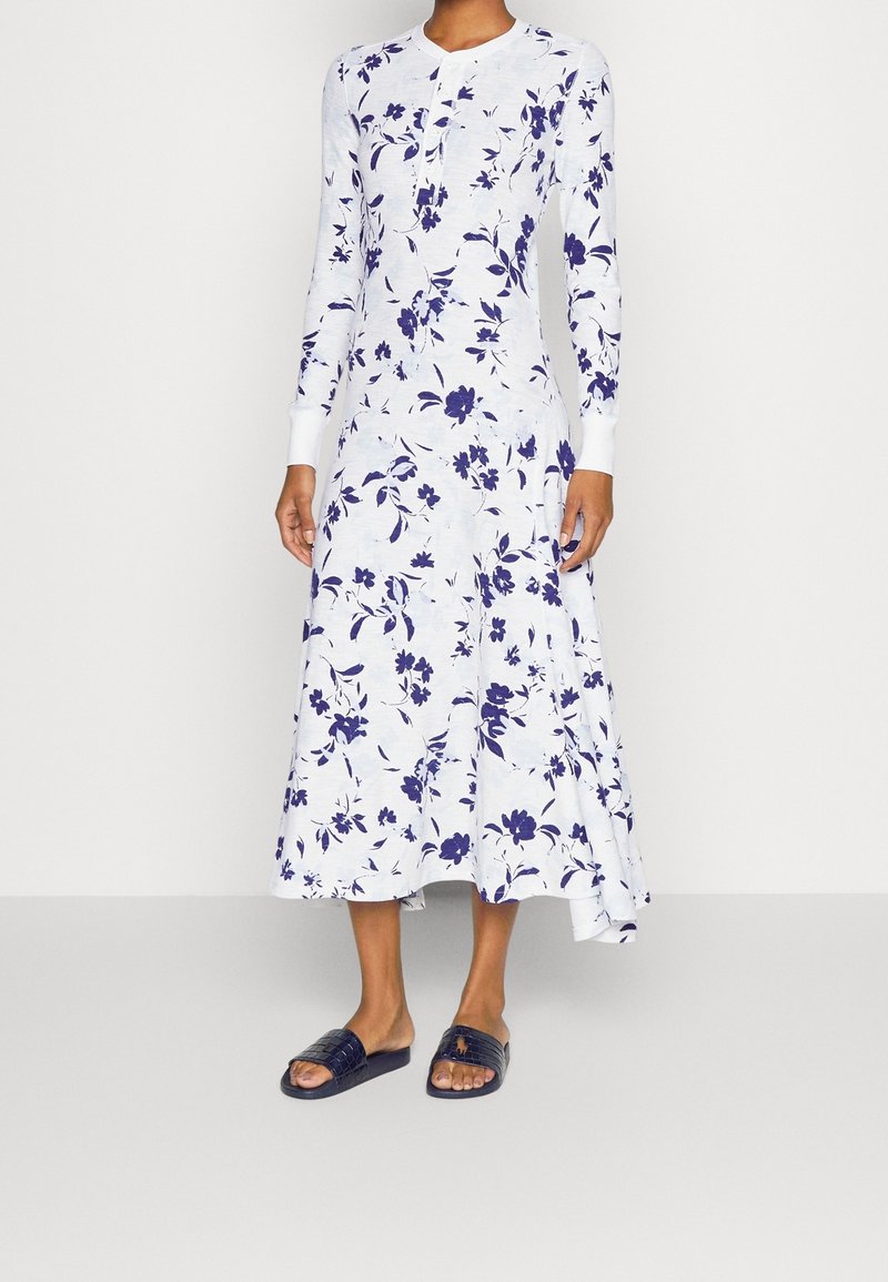 Robe blanche à manches longues avec imprimé floral violet, corsage ajusté, jupe évasée et poignets côtelés. Associée à des sandales noires à enfiler.