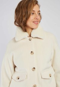 Kremowy wełniany płaszcz z puszystym białym kołnierzem, ozdobiony dużymi guzikami i dwoma bocznymi kieszeniami. Miękka tekstura, luźny krój i minimalistyczny styl.