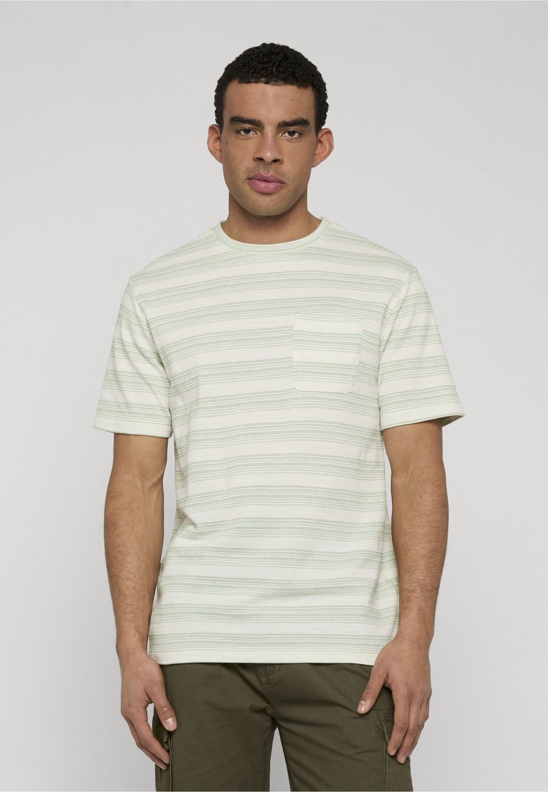 Homme portant un T-shirt à manches courtes rayé vert clair et blanc avec une poche poitrine et un pantalon vert olive, debout devant un fond uni.