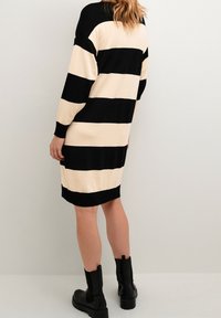Robe pull rayée noire et crème avec des manches longues, coupe droite, poignets côtelés et longueur mi-cuisse, associée à des bottines noires.