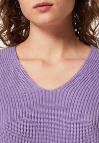 Suéter de punto lila con cuello en V, textura acanalada, patrón uniforme y un corte relajado que se detiene por encima de la clavícula.