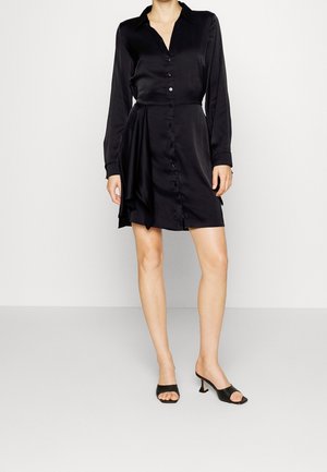 Kvinde iført en sort satin kjole med knapper, lange ærmer og asymmetrisk draperet detalje, kombineret med sorte åbne tåhælede sandaler.