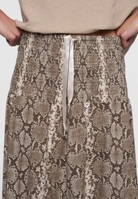 Bruine rok met slangenprint en een gestructureerde afwerking. Is voorzien van een smockband aan de taille en een satijnen trekkoord met metalen aglets voor aanpassing.