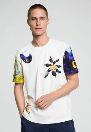 Mann mit kurzen Haaren trägt ein weißes T-Shirt mit bunten abstrakten Mustern auf Ärmeln, Brust und unterem linken Saum, steht vor einem grauen Hintergrund.