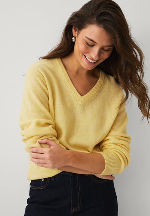 Vrouw met lang bruin haar, zilveren oorringen en een gele v-trui, glimlachend en met gekruiste armen.