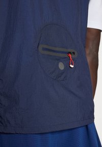 Gros plan sur une poche de veste marine avec une fermeture éclair horizontale et un cordon rouge, portée sur un pantalon bleu et une manche blanche visible.