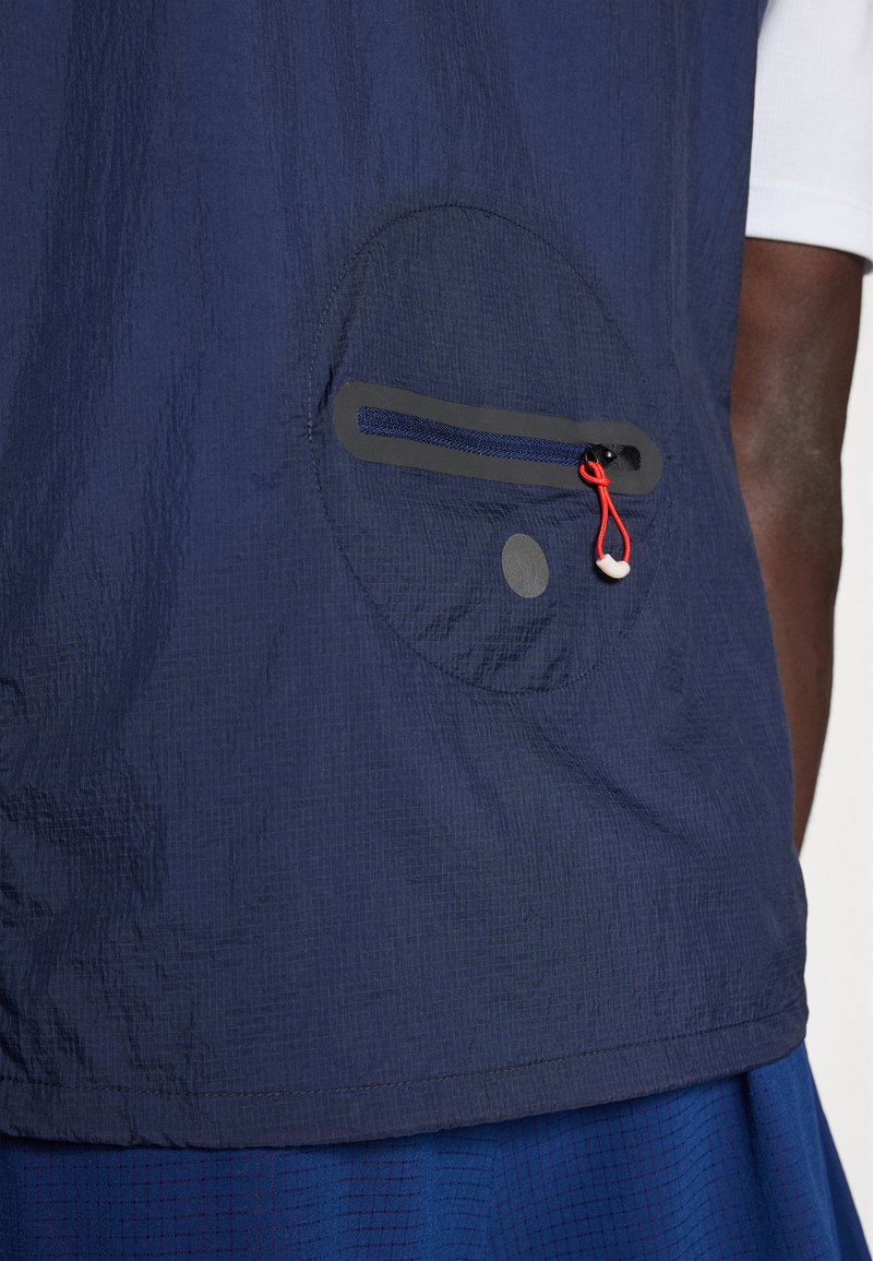 Gros plan sur une poche de veste marine avec une fermeture éclair horizontale et un cordon rouge, portée sur un pantalon bleu et une manche blanche visible.