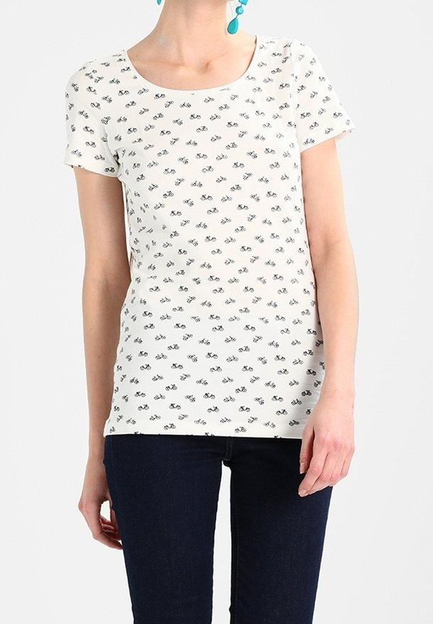 Femme portant une chemise blanche à manches courtes avec de petits imprimés noirs de bicyclettes et un jean bleu foncé, montrée du cou aux hanches.