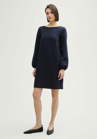 Abito a maniche lunghe blu navy con scollo a barca, silhouette dritta e polsini elasticizzati. Abbinato a scarpe nere flat.