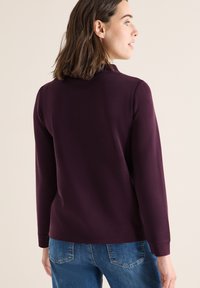 Langärmliger Pullover in tiefem Lila, mit glatter Textur und lockerer Passform, kombiniert mit hellblauen Jeans, von hinten betrachtet.