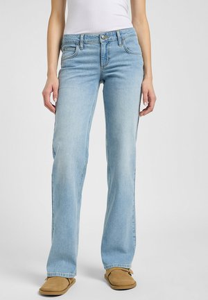 Woman wearing light blue straight-leg jeans, white ribbed tank top, and tan slip-on shoes, standing against plain background.