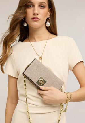 Kvinde i en cremefarvet kjole, der holder en glitrende guldfarvet clutch med kædestrop, iført perledråbeørenringe, en gylden halskæde og armbånd.