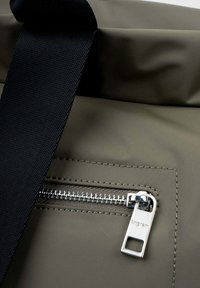 Sac olive en matière texturée et résistante à l'eau. Il dispose d'une poche zippée argentée et d'une sangle en sangle noire pour le transport.