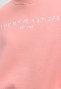 Ροζ φούτερ με λευκό λογότυπο Tommy Hilfiger και κείμενο "EST. 1985" τυπωμένα στο στήθος κάτω από έναν ψηλό λαιμό με στρογγυλή λαιμόκοψη.