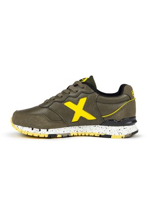 Zapatilla deportiva de cuero verde oliva con un logo X amarillo. Presenta una suela moteada en blanco y amarillo y acentos de malla negra en el collar.