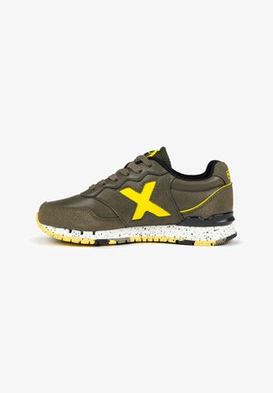 Zapatilla deportiva de cuero verde oliva con un logo X amarillo. Presenta una suela moteada en blanco y amarillo y acentos de malla negra en el collar.