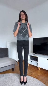 Grå stickad tröja med korta ärmar och diamantmönster, kombinerad med svarta tajta jeans med dekorativa detaljer på sidorna och svarta klackar.