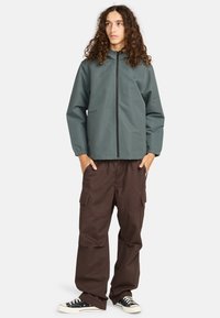 Junge Person mit langen, lockigen Haaren, die eine graue Zip-Jacke, braune Cargo-Hosen und schwarze Sneaker trägt, steht mit den Händen in den Taschen.