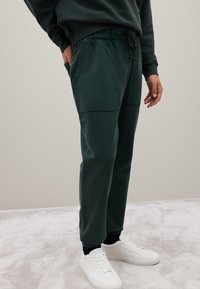 Donkergroene sweatpants met een elastische tailleband en trekkoord, met twee voorzakken en taps toelopende boorden. Draagt met witte sneakers.