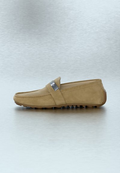 Ανδρικό μοκασίνι από μπεζ σουέντ με λαστιχένια σόλα και ασημένια μεταλλική πινακίδα στο πλάι, εμφανιζόμενο σε προφίλ σε μια ανακλαστική επιφάνεια.