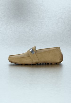 Mocassin pour homme en daim beige avec semelle en caoutchouc antidérapante et étiquette métallique argentée sur le côté, présenté de profil sur une surface réfléchissante.