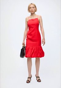 Femme aux cheveux blonds courts portant une robe rouge ajustée avec des bretelles fines et un ourlet à volants, tenant un sac à main noir et portant des sandales noires à talons hauts.