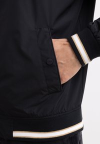 Juodas bomberio stiliaus švarkas su priekinėmis kišenėmis, pasižymintis šonkaulinėmis rankovėmis su baltomis ir auksinėmis juostelėmis, bei elastingu juosmeniu su kontrastingomis detalėmis.