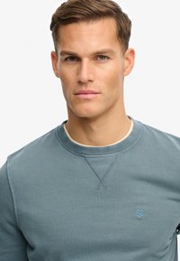 Sudadera azul con cuello redondo y textura suave, que presenta un pequeño emblema en el pecho izquierdo y un ribete de contraste en el cuello.